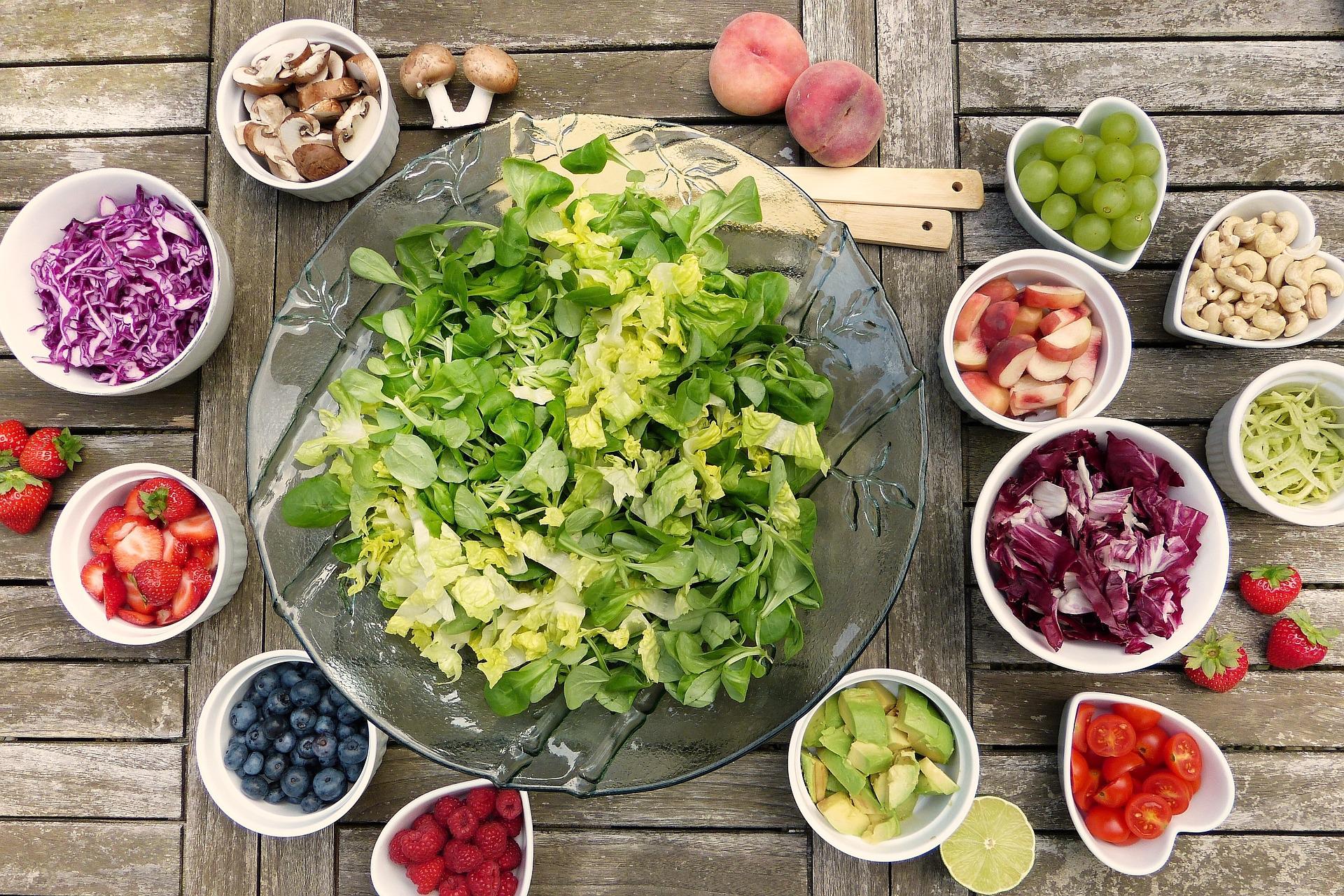 Ein grüner Salat steht in einer farblosen Glasschale in der Bildmitte, ringsherum in kleineren weißen Schalen befinden sich sommerliche Zutaten, z.B. Beeren