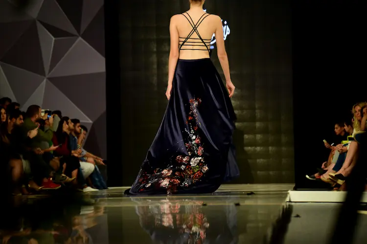 Eine Modenschau, das Model trägt ein schwarzes Kleid und man sieht ihren Rücken, an der Seite Zuschauer